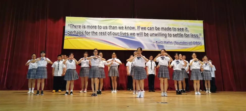 Students in school uniforms perform on a stage with a Kurt Hahn quote banner behind them.